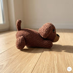 A brown crocheted plush dog toy lying on a light wood floor in a sunlit room.