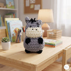 A crocheted plush toy resembling a hippo or donkey sits on a light wood table in a cozy, softly lit room. The toy is made of gray yarn with a cream-colored snout and mouth area, black ears, hooves, and hair, and small black embroidered eyes.