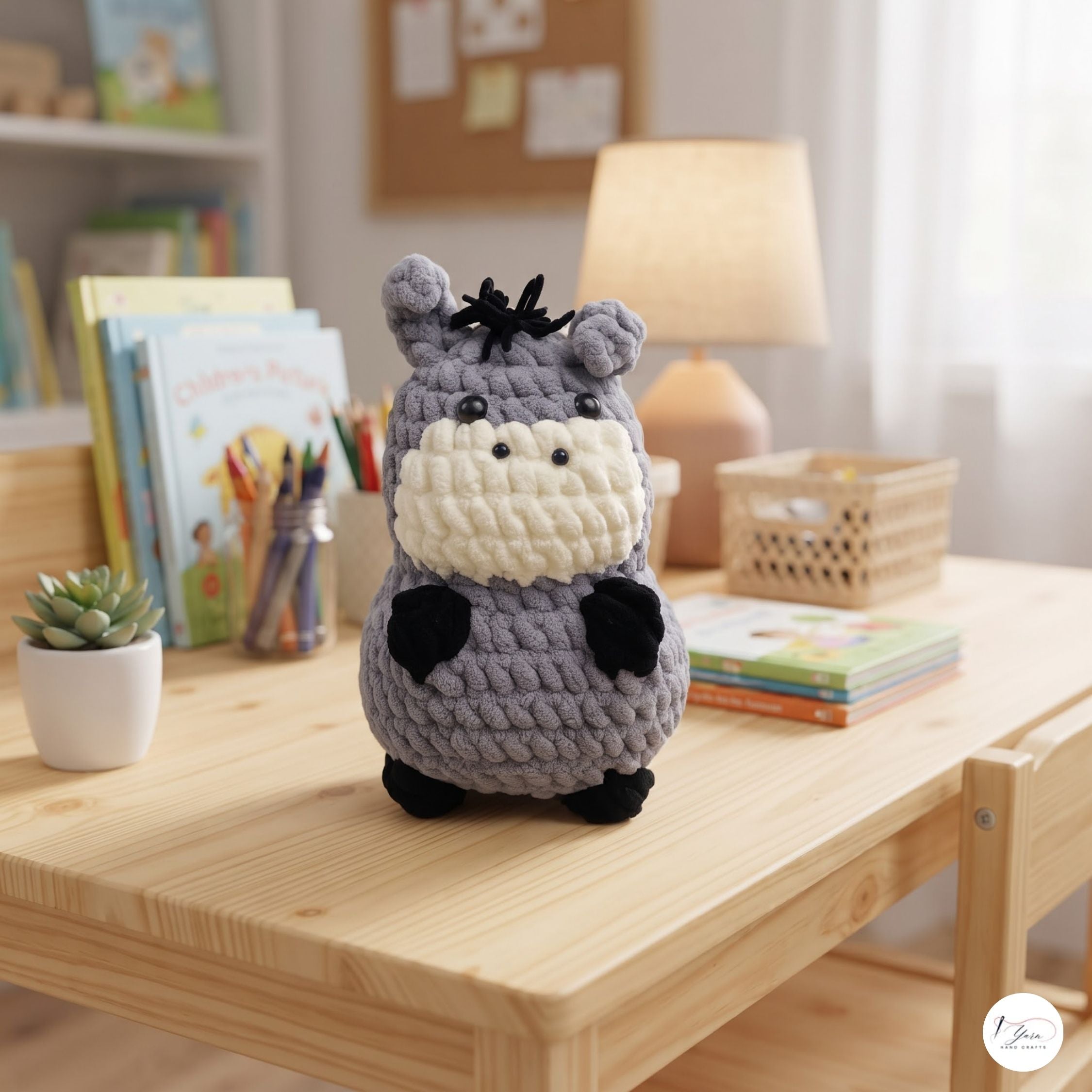 A crocheted plush toy resembling a hippo or donkey sits on a light wood table in a cozy, softly lit room. The toy is made of gray yarn with a cream-colored snout and mouth area, black ears, hooves, and hair, and small black embroidered eyes.