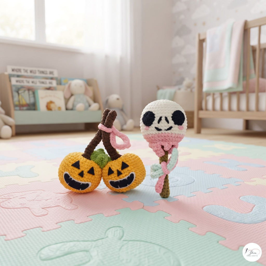 Two handmade crocheted toys displayed on a pastel puzzle playmat in a softly lit nursery. One toy features two orange jack-o'-lantern heads on brown stems with pink loops, and the other is a white skull holding a pink and green flower stem.