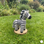 A handmade crocheted zebra toy standing on a round wooden slice base, placed on a green grass lawn with a garden background.