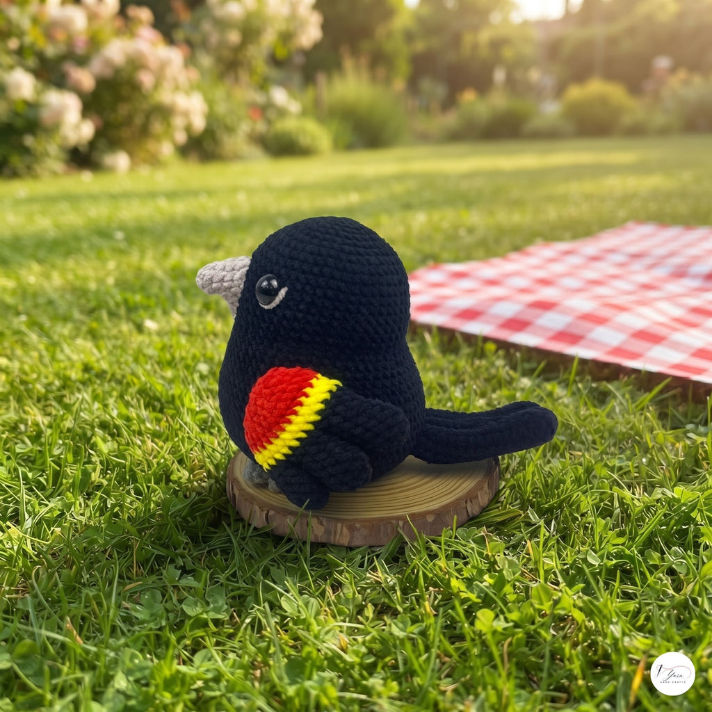 A crocheted bird toy sits on a small wooden slice on a grassy lawn, with a red-and-white checkered blanket in the background and a blurred garden scene behind it.