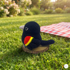 A crocheted bird toy sits on a small wooden slice on a grassy lawn, with a red-and-white checkered blanket in the background and a blurred garden scene behind it.