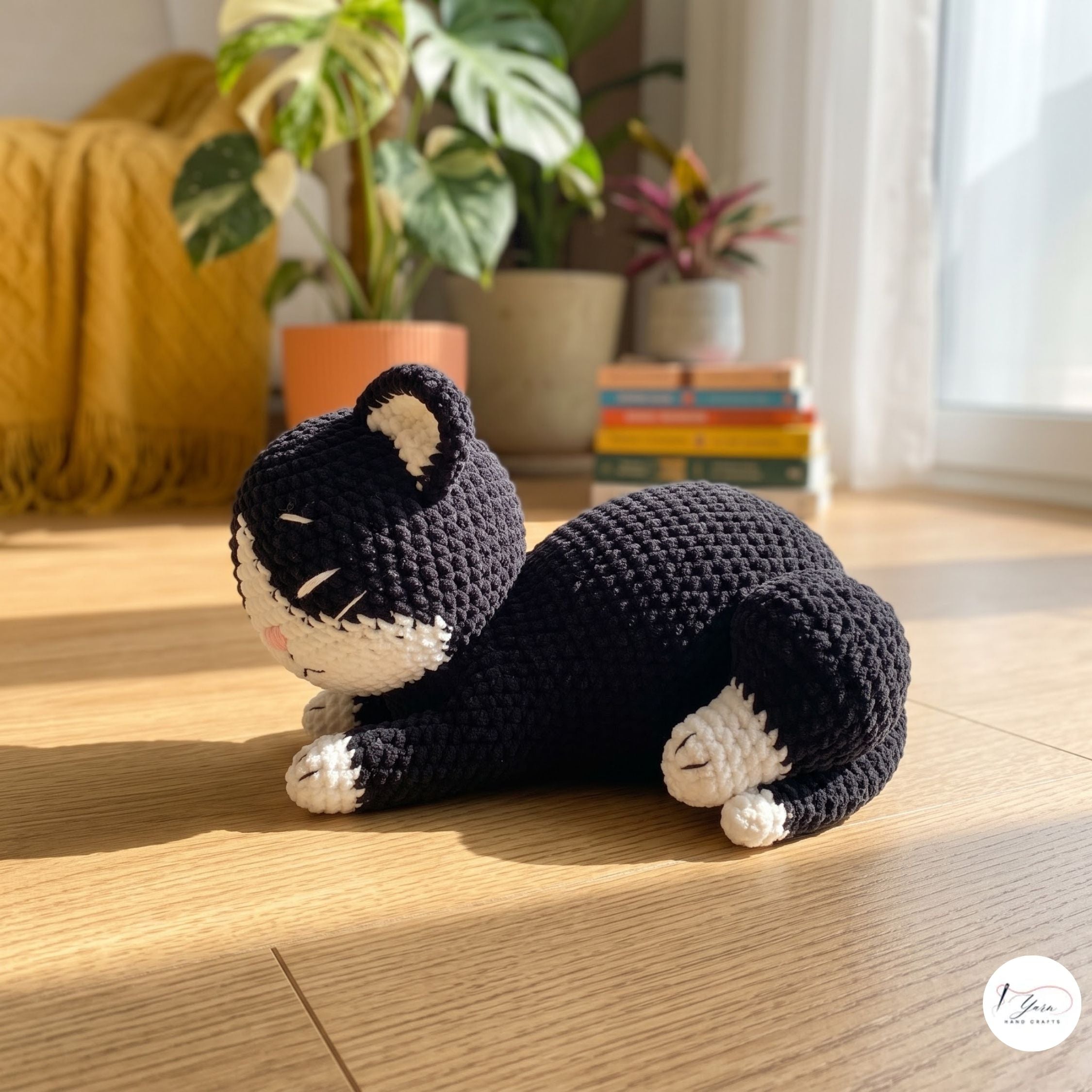 A handmade crocheted black and white cat toy lying on a wooden floor in a bright, sunlit room with potted plants and a yellow blanket in the background.