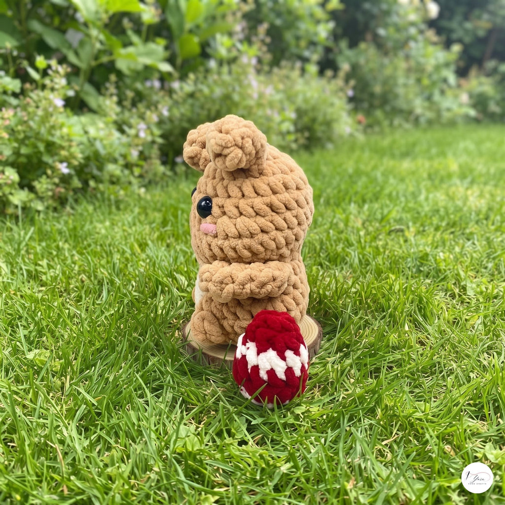 A small crocheted bunny toy sits on green grass outdoors, with a red and white crocheted egg in front of it and a blurred garden background.