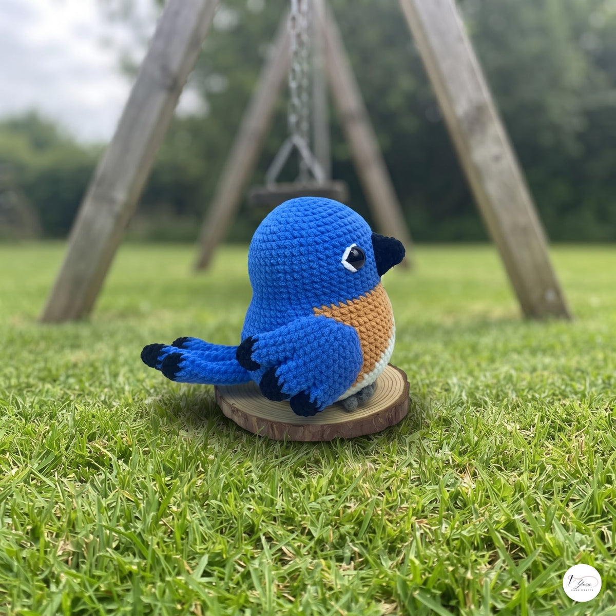 A crocheted blue bird toy with a tan belly, black beak and feet, sitting on a round wooden slice base on a green grass lawn with a blurred wooden swing in the background.