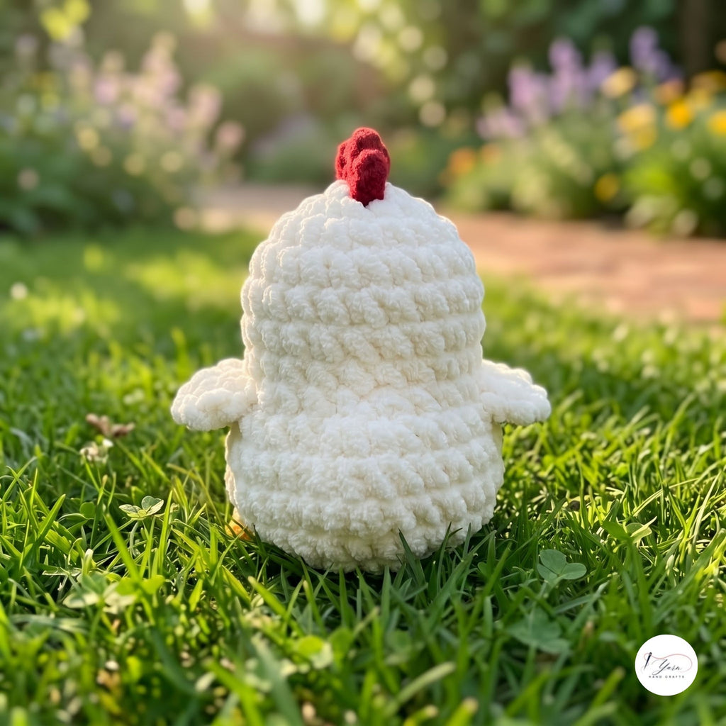 A crocheted white chicken toy with a red comb sits on green grass in a sunny garden with a blurred background of flowers and foliage.