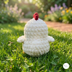 A crocheted white chicken toy with a red comb sits on green grass in a sunny garden with a blurred background of flowers and foliage.