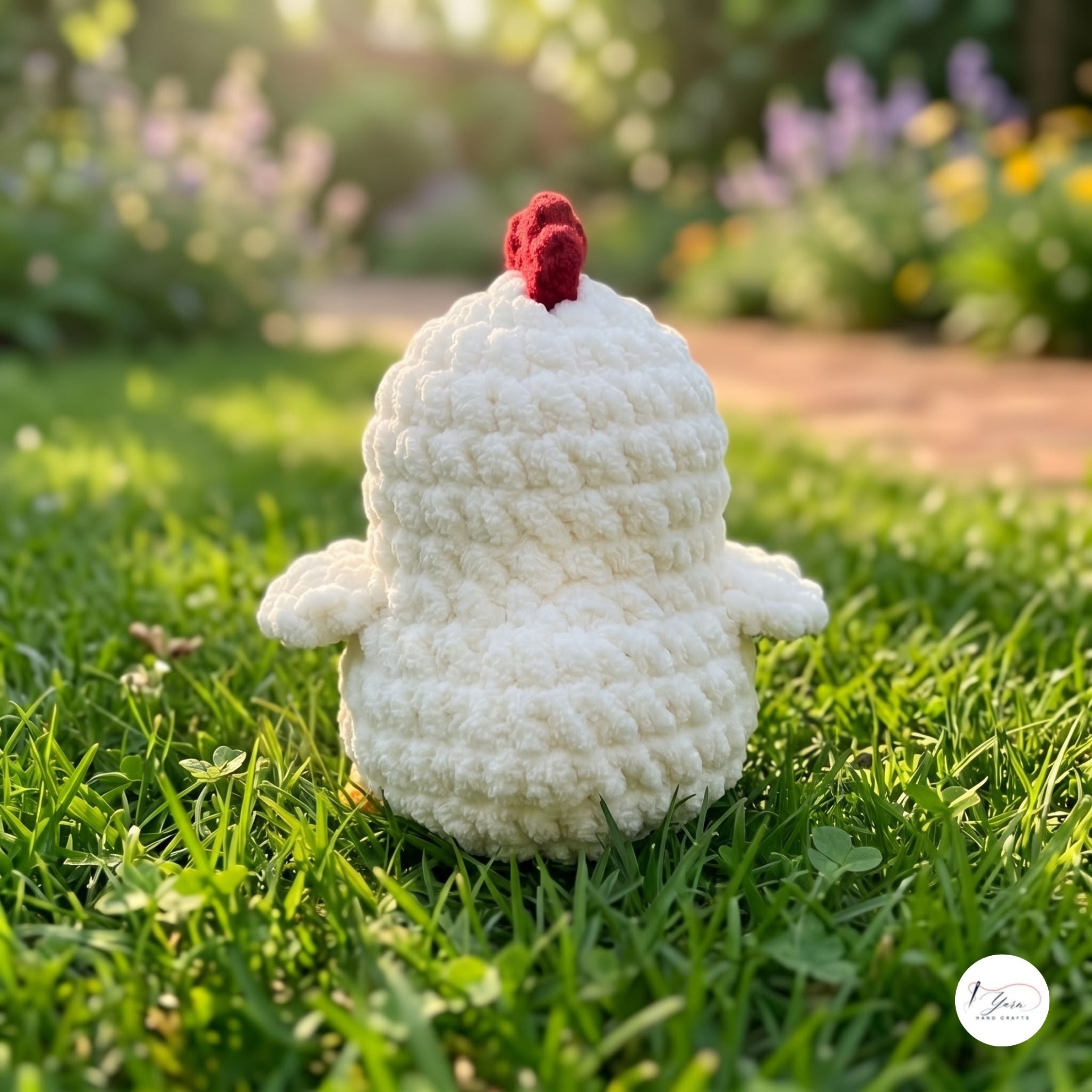 A crocheted white chicken toy with a red comb sits on green grass in a sunny garden with a blurred background of flowers and foliage.