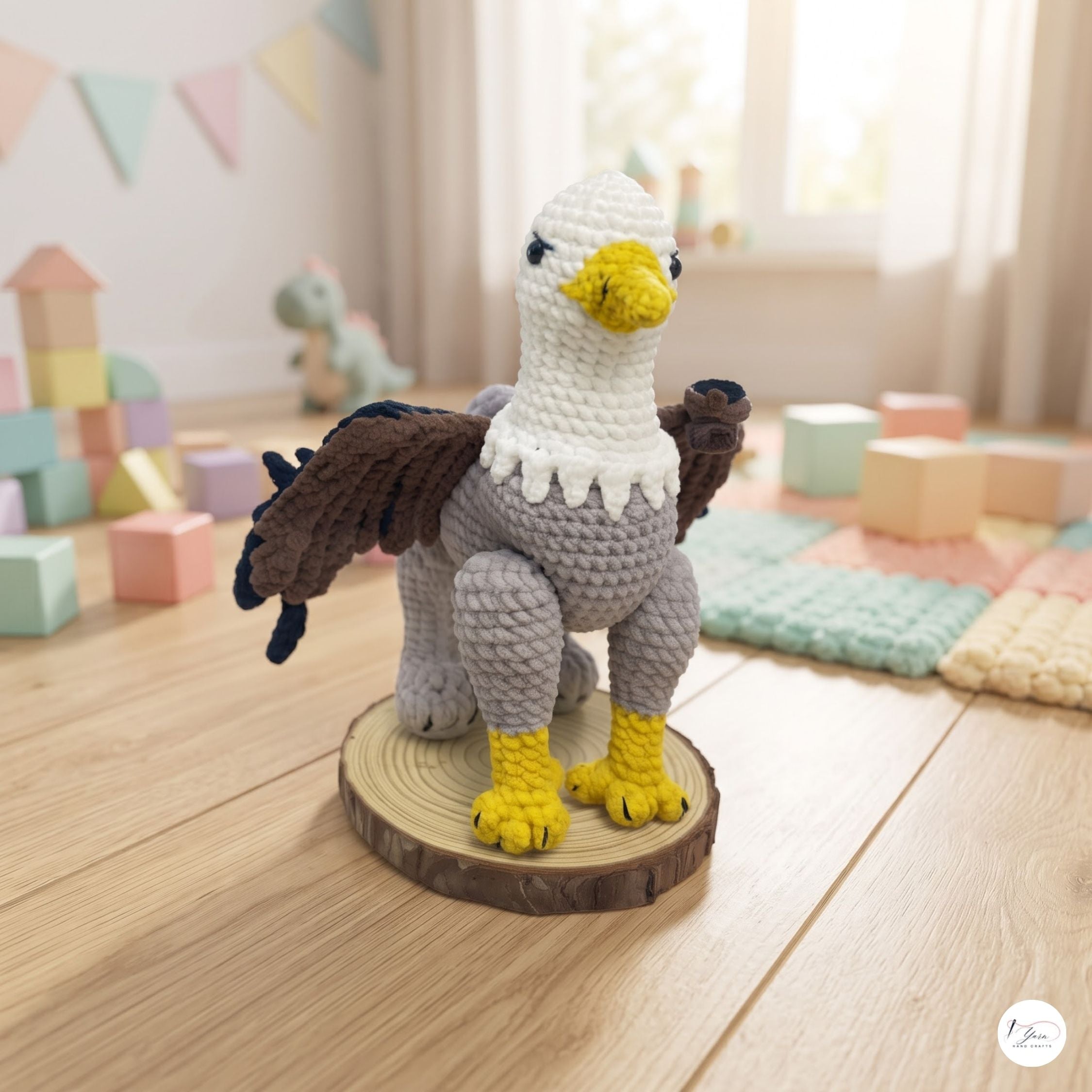 A crocheted eagle toy standing on a round wood slice base, photographed in a softly lit children's room with pastel blocks and toys blurred in the background.