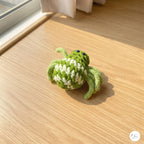 A small crocheted spider toy in light green and white sits on a light wood floor near a window, with warm natural sunlight casting a soft shadow.