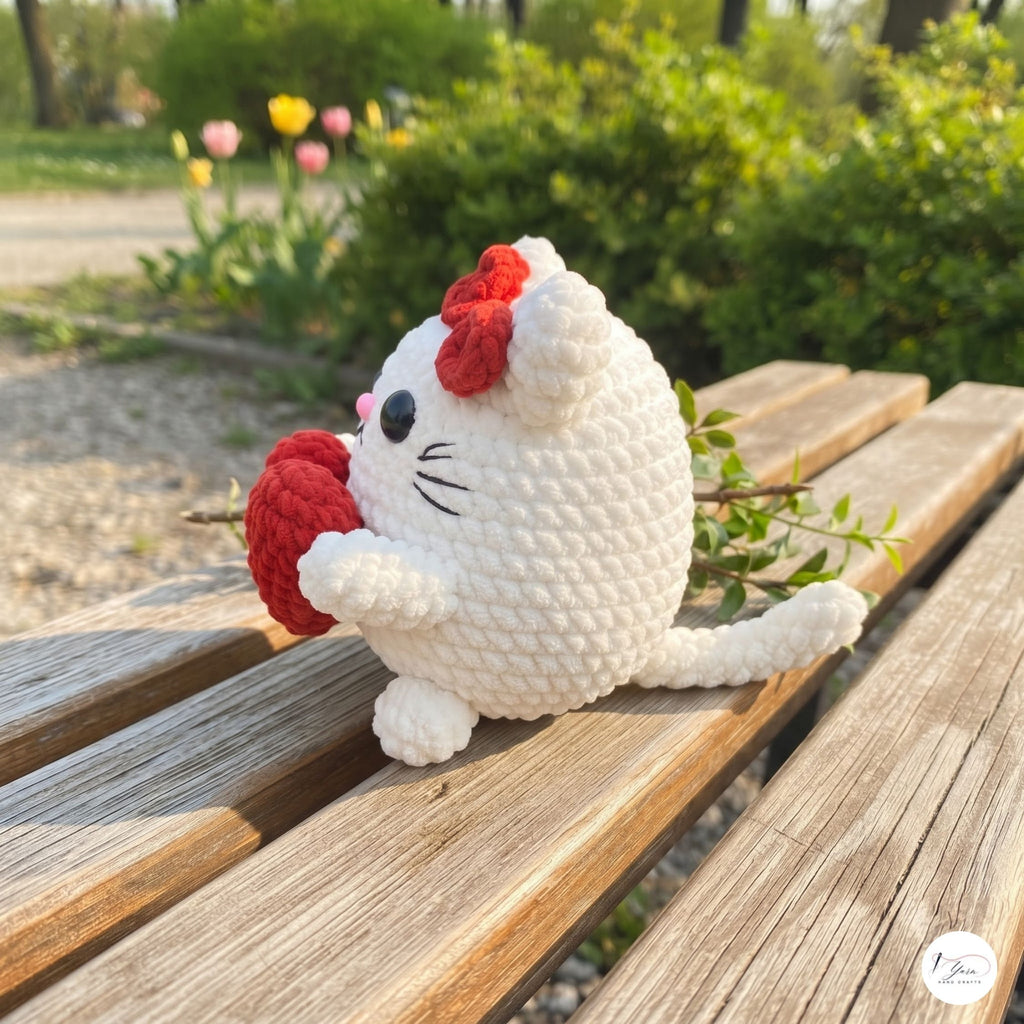 A white crocheted cat plush toy with a red bow and red heart-shaped accessory, sitting on a wooden bench in an outdoor garden setting.