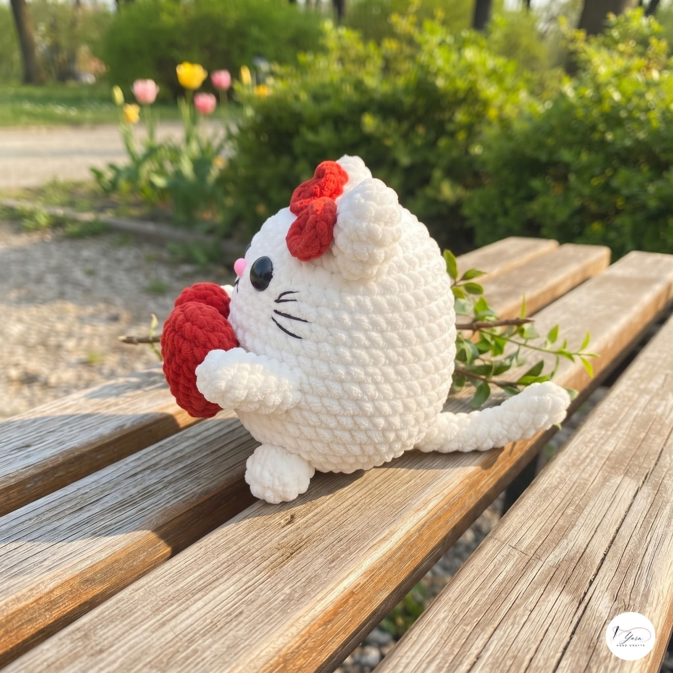 A white crocheted cat plush toy with a red bow and red heart-shaped accessory, sitting on a wooden bench in an outdoor garden setting.