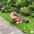 A crocheted amigurumi tiger toy sitting on a green grassy lawn next to a stone path, with a blurred garden background.