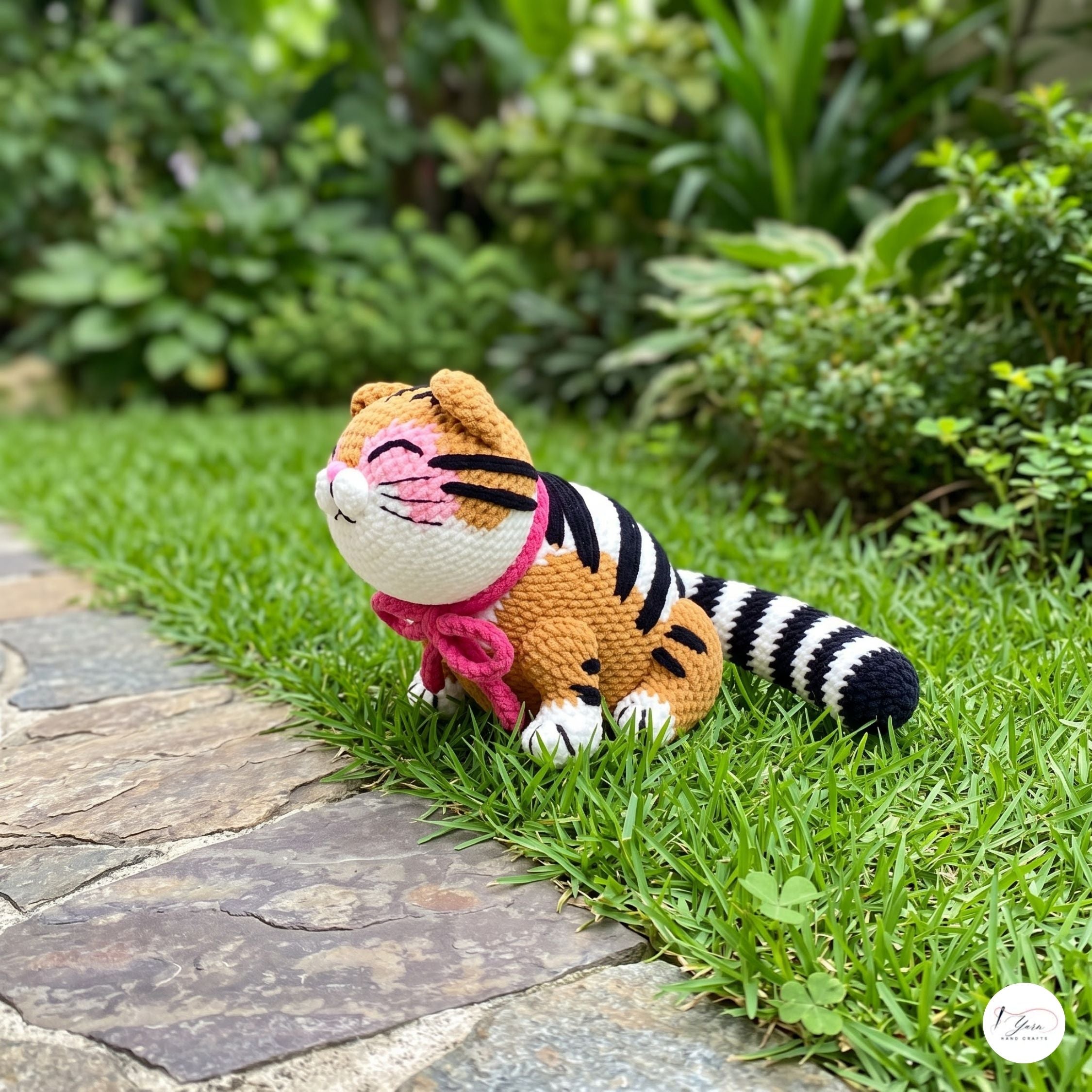 A crocheted amigurumi tiger toy sitting on a green grassy lawn next to a stone path, with a blurred garden background.