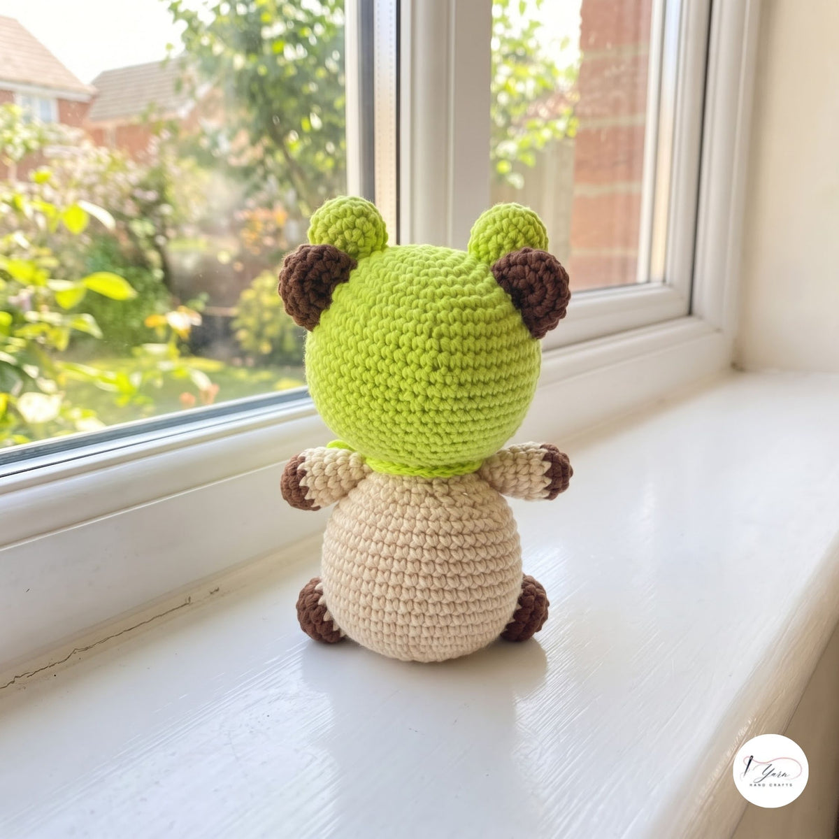 A crocheted plush toy frog sitting on a white windowsill, looking out towards a garden. The toy has a lime green head and back, a cream-colored belly, and dark brown ears, paws, and feet.