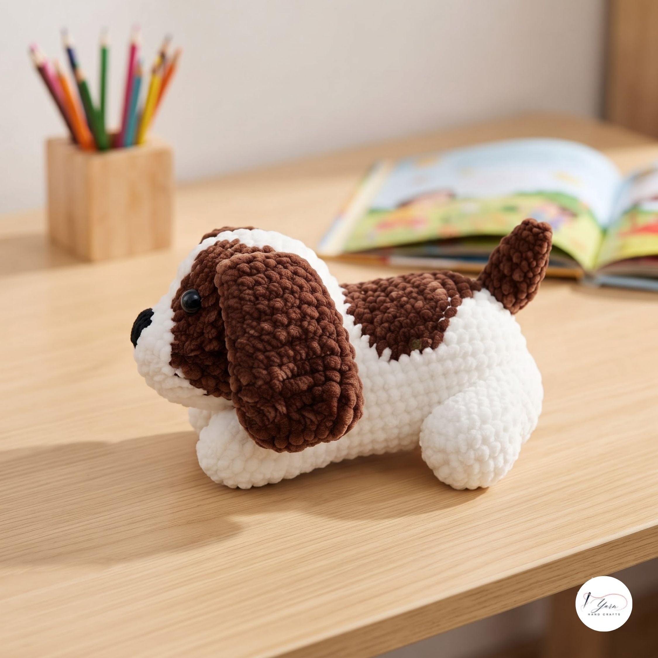 A crocheted dog toy with brown and white textured yarn sits on a light wood desk, with a pencil holder and an open book blurred in the background.