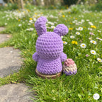 A purple crocheted bunny toy with long ears sits on a small wooden stand in a grassy garden with white and yellow daisies.