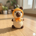 A crocheted plush toy shaped like a bear wearing an orange pumpkin hat and matching orange scarf, sitting on a wooden floor in a softly blurred indoor setting.