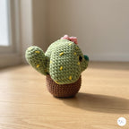 A side view of a plush handmade crochet cactus puppy toy with floppy ears, yellow embroidered spines, and a pink flower on top, sitting in a brown crochet pot base on a sunlit wooden floor.