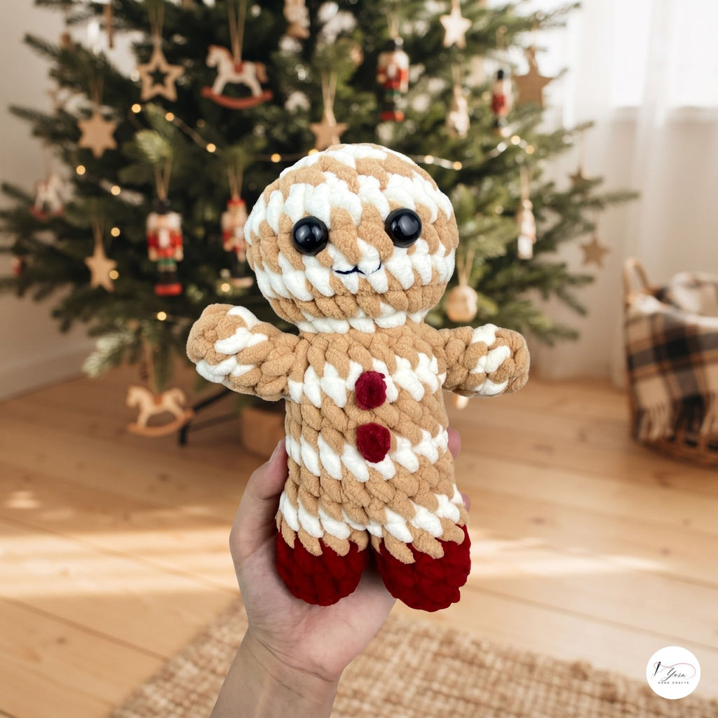 A handmade crocheted gingerbread person doll held in a hand, set against a blurred background of a decorated Christmas tree and warm indoor room.