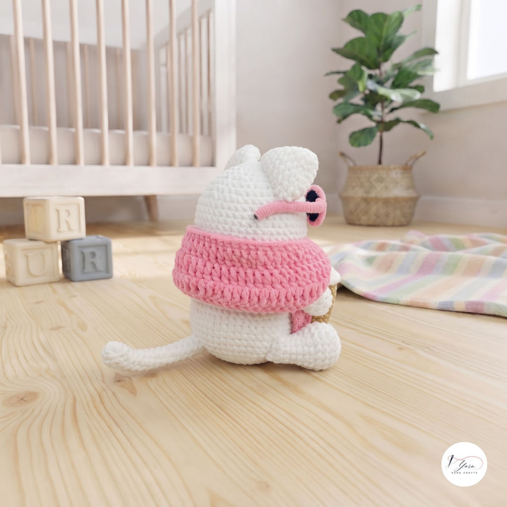 A side view of a cute handmade white crochet cat plush sitting on a light wooden floor in a cozy nursery setting, wearing a pink sweater, pink sunglasses resting over its eyes, and showing its white tail extended behind.