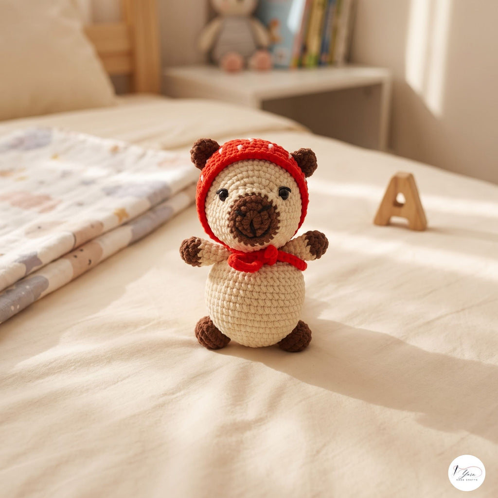 A crocheted teddy bear toy sits on a cream-colored bed in a softly lit bedroom. The bear has a cream body, brown paws and snout, and wears a red knit hat and matching red scarf.