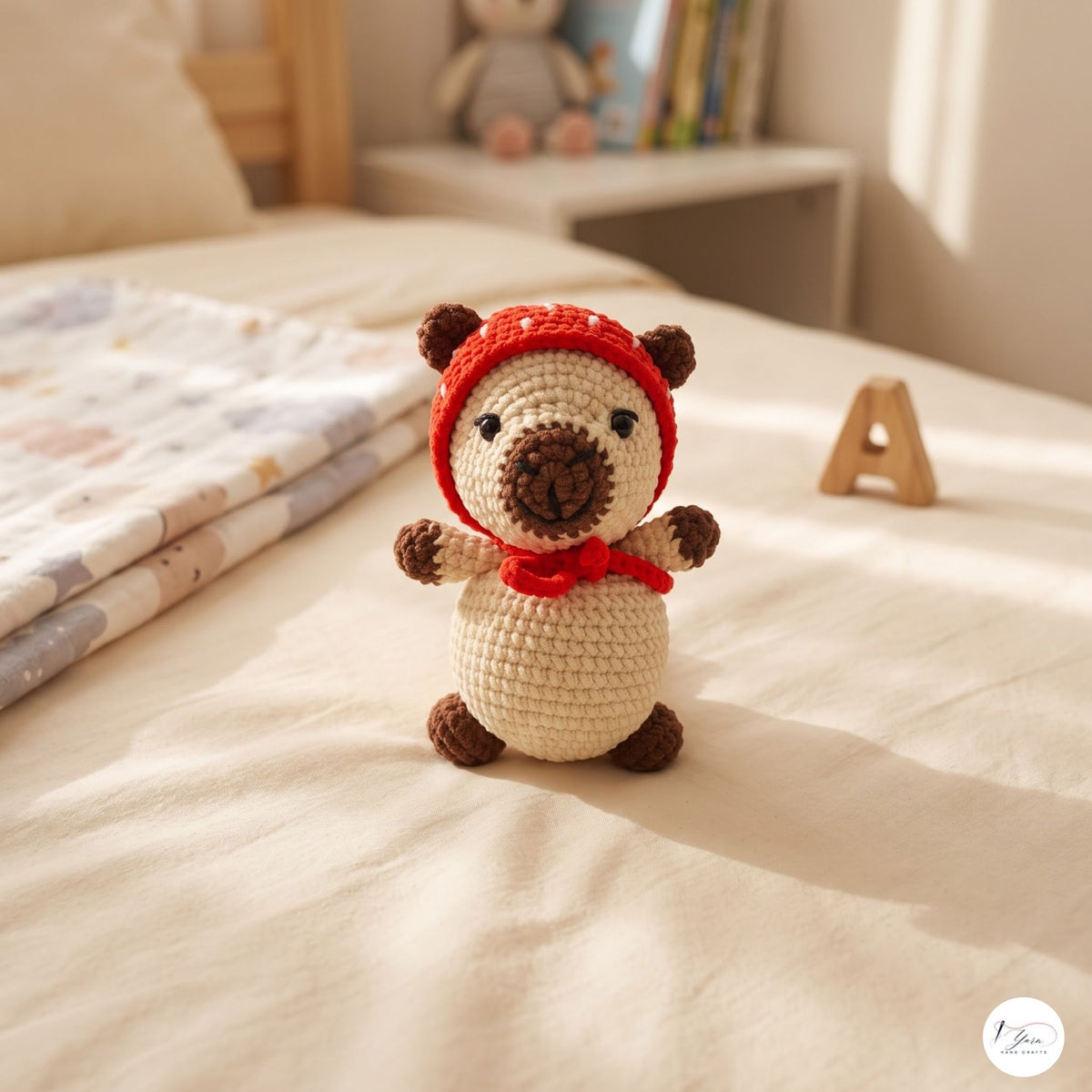 A crocheted teddy bear toy sits on a cream-colored bed in a softly lit bedroom. The bear has a cream body, brown paws and snout, and wears a red knit hat and matching red scarf.