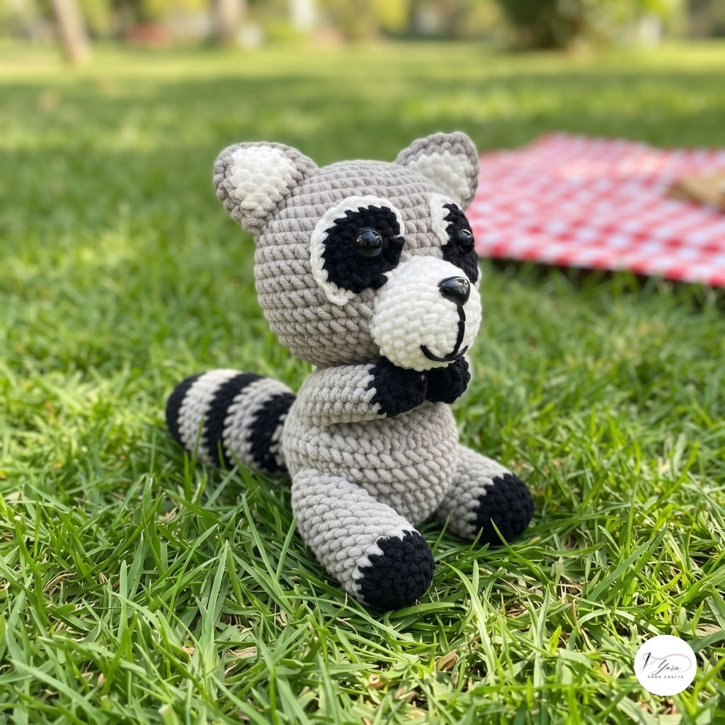 A crocheted raccoon toy sitting on green grass outdoors, with a blurred red-and-white picnic blanket in the background.