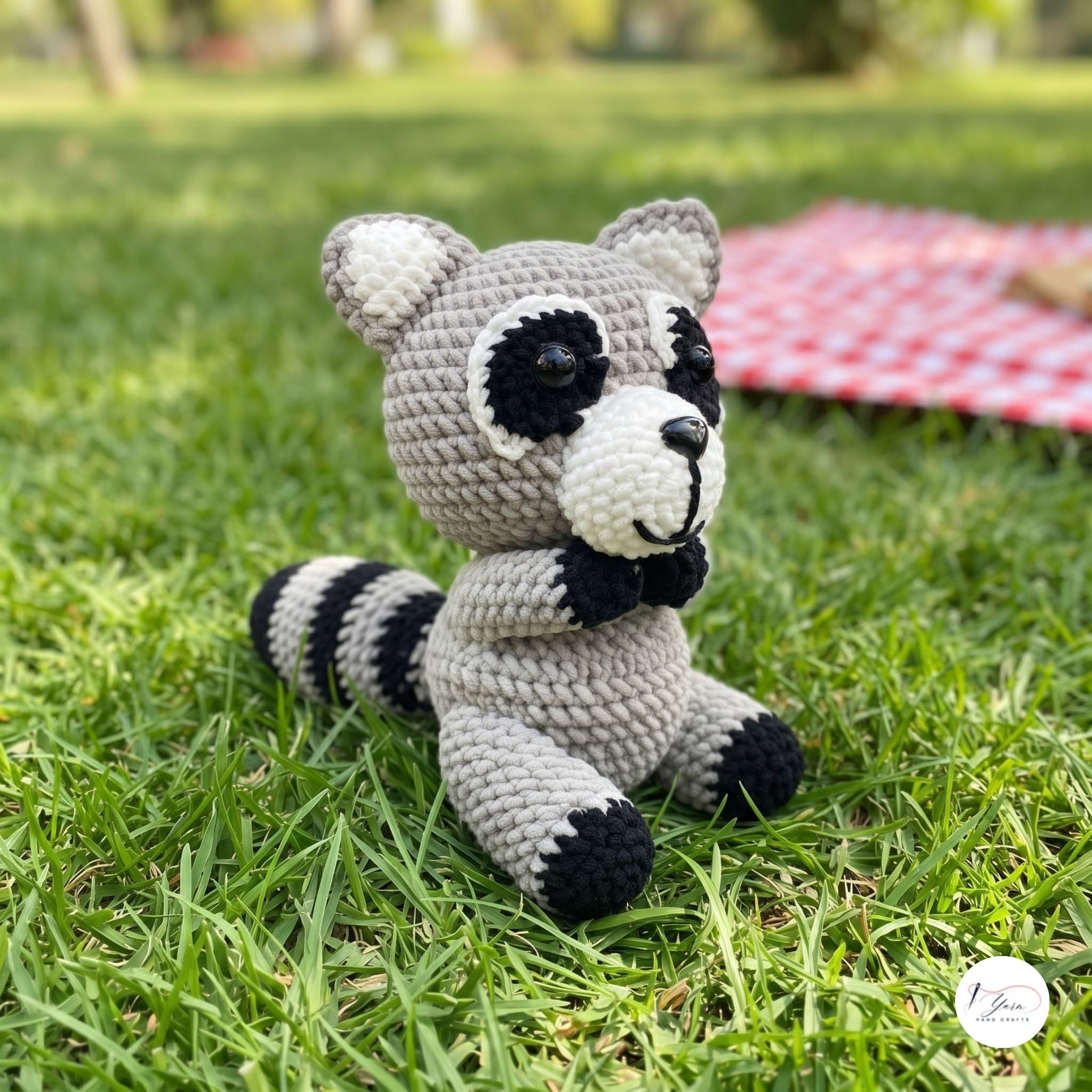 A crocheted raccoon toy sitting on green grass outdoors, with a blurred red-and-white picnic blanket in the background.