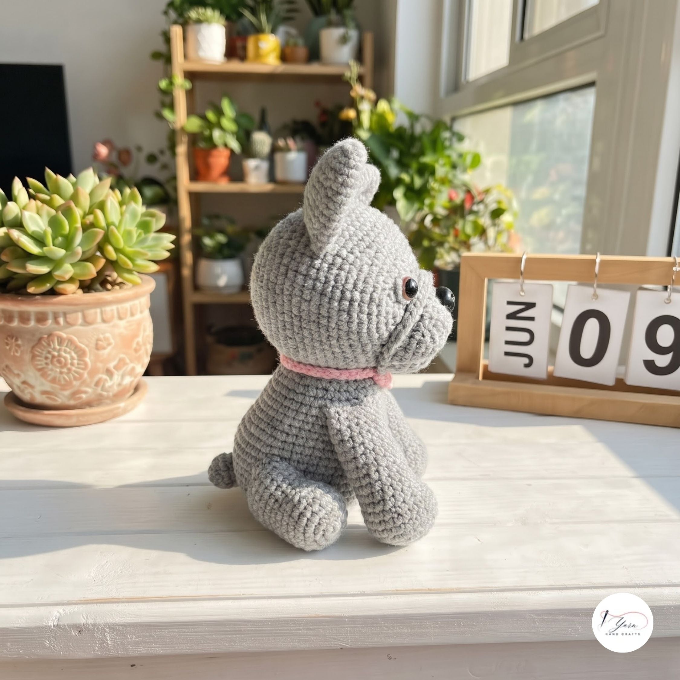 A gray crocheted dog toy with a pink collar sits on a white windowsill in a bright room with potted plants and a wooden shelf in the background.