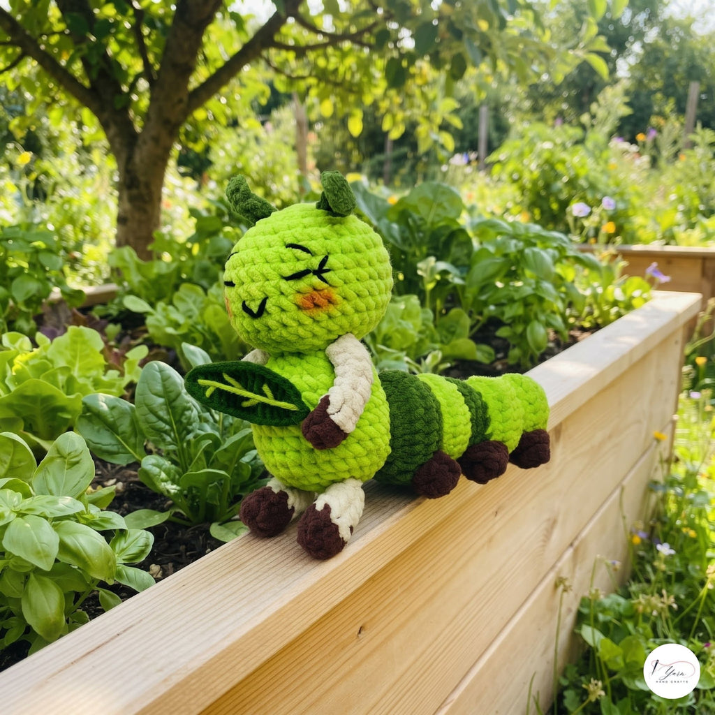 A handmade crocheted caterpillar toy in shades of green, cream, and brown, holding a leaf and sitting on a wooden garden bed with a lush garden background.