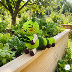 A handmade crocheted caterpillar toy in shades of green, cream, and brown, holding a leaf and sitting on a wooden garden bed with a lush garden background.