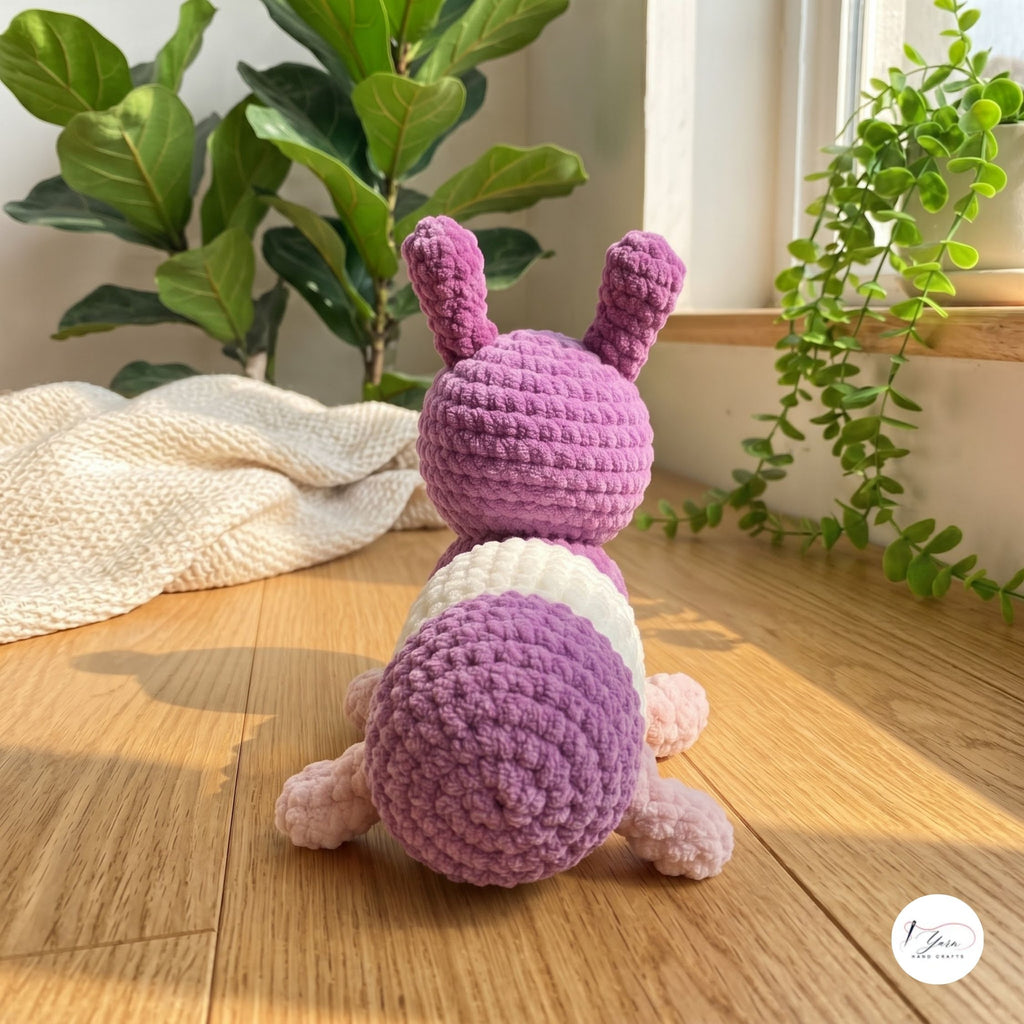 A crocheted bunny toy shown from the back, featuring a purple body, white belly, and pink legs, set on a wooden floor with houseplants in the background.