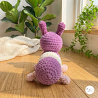 A crocheted bunny toy shown from the back, featuring a purple body, white belly, and pink legs, set on a wooden floor with houseplants in the background.
