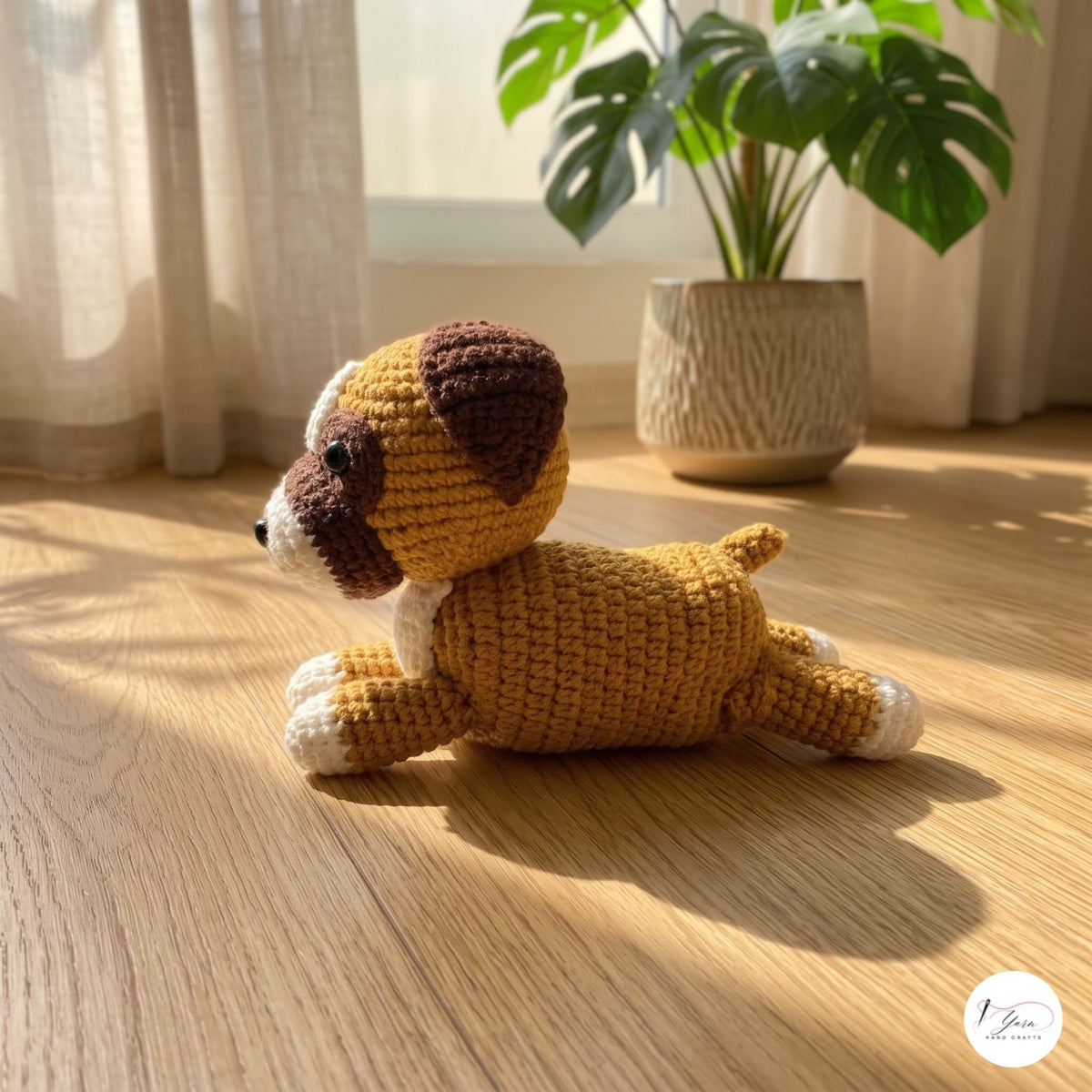 A crocheted plush puppy toy in mustard yellow, brown, and cream, lying on a sunlit wooden floor with a potted plant and sheer curtains in the background.