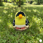 A handmade crocheted bird figurine in yellow, black, white, and orange, standing on a small wooden base in a grassy outdoor setting.