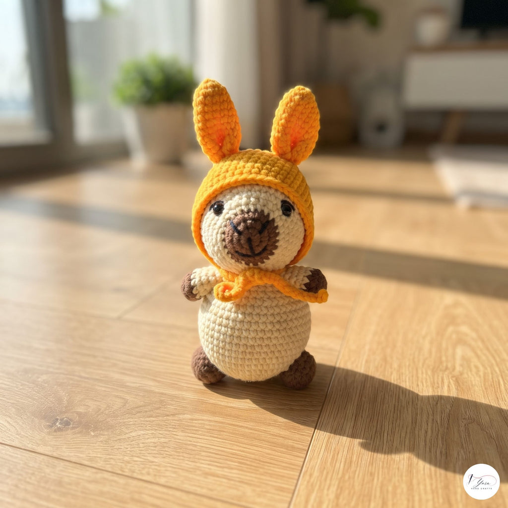 A crocheted kangaroo toy standing on a light wooden floor in a sunlit room. The toy has a cream body, brown snout and feet, and a yellow-orange hood with long ears and a matching neck scarf.