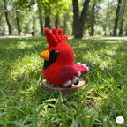 A crocheted cardinal bird toy in red, black, yellow, and pink sits on a small round wooden stand on a grassy lawn in a sunny park.