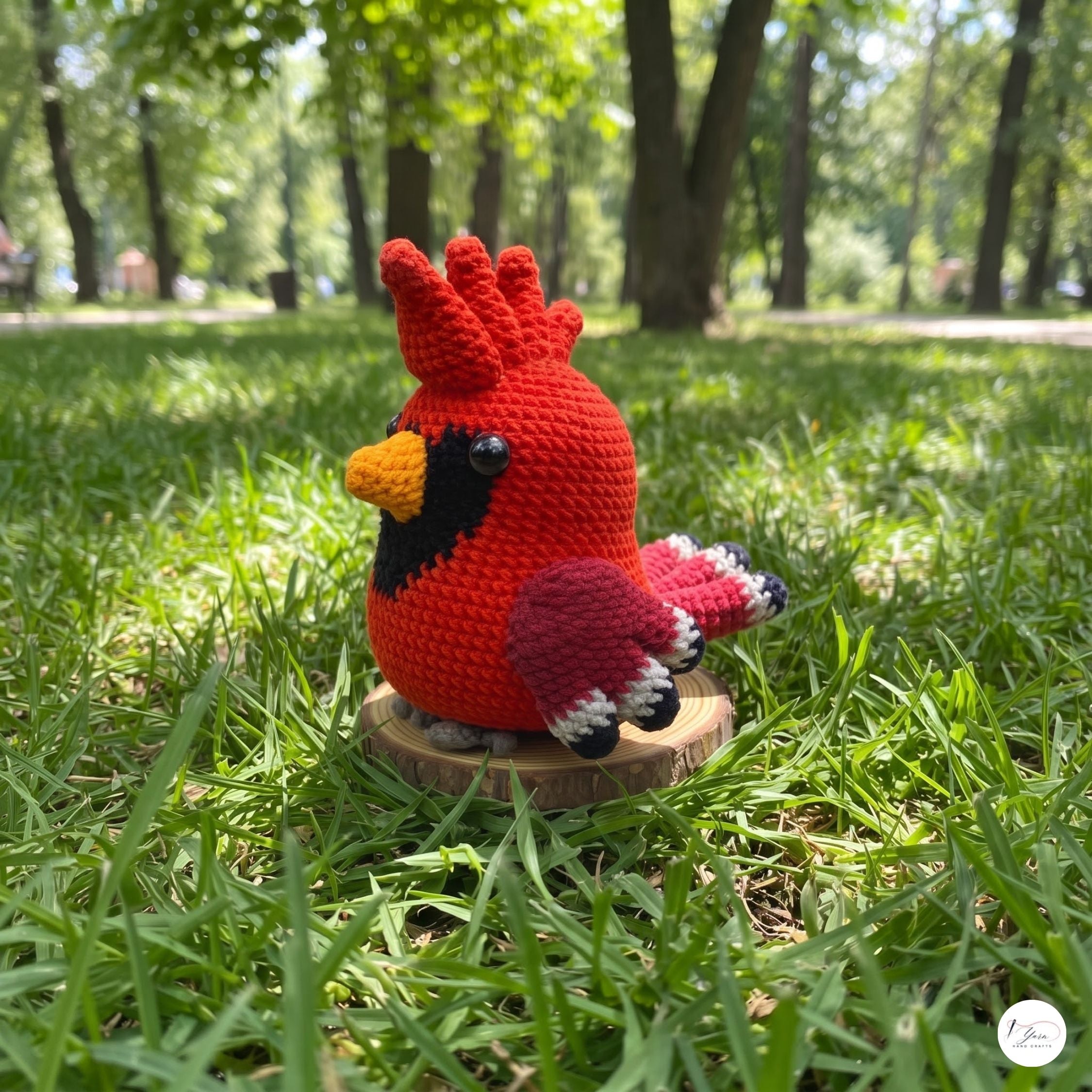 A crocheted cardinal bird toy in red, black, yellow, and pink sits on a small round wooden stand on a grassy lawn in a sunny park.