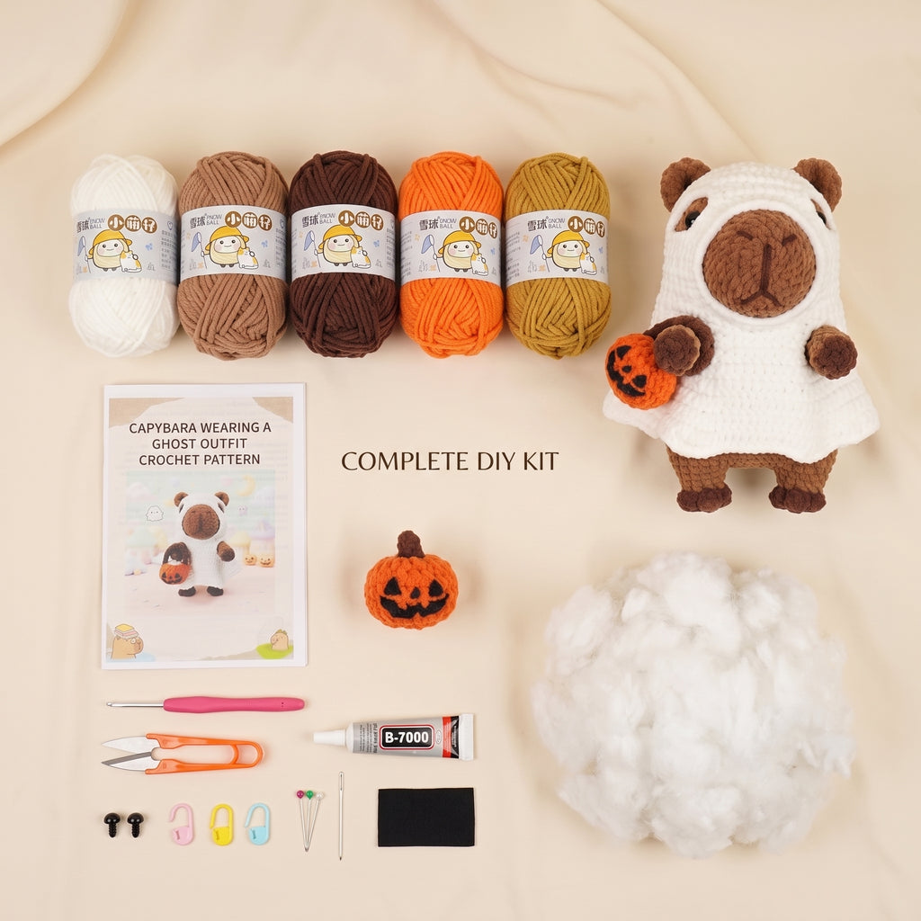 A DIY crochet kit arranged on a beige fabric background, including four yarn balls in white, tan, brown, orange, and mustard, a capybara wearing a ghost outfit finished product, a printed pattern booklet, a small orange pumpkin, white stuffing, and crochet tools.