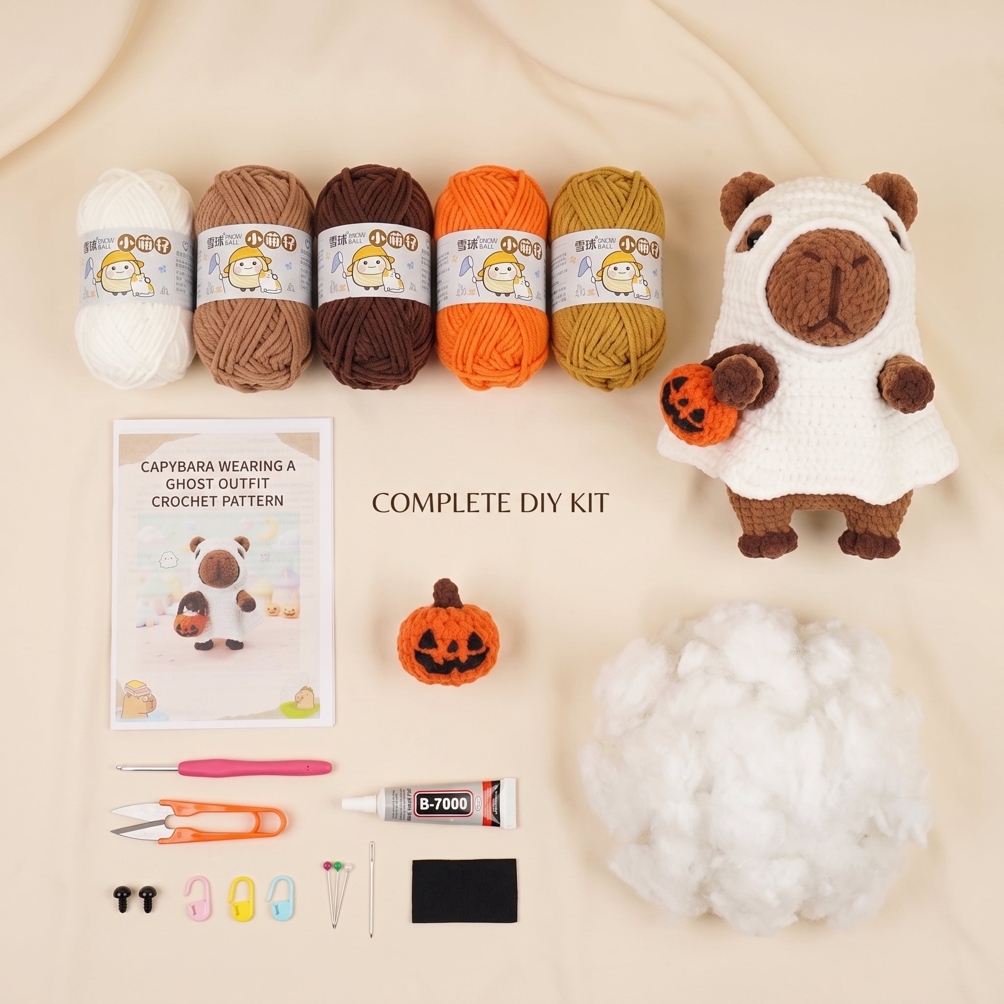 A DIY crochet kit arranged on a beige fabric background, including four yarn balls in white, tan, brown, orange, and mustard, a capybara wearing a ghost outfit finished product, a printed pattern booklet, a small orange pumpkin, white stuffing, and crochet tools.