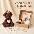 Cookie Puppy Crochet Kit displayed on a beige fabric backdrop, featuring a crocheted brown puppy with a white heart, yarn balls, a crochet hook, a pattern booklet, and small cookie pieces arranged beside an open kraft box with the text 'COOKIE PUPPY CROCHET KIT' above.