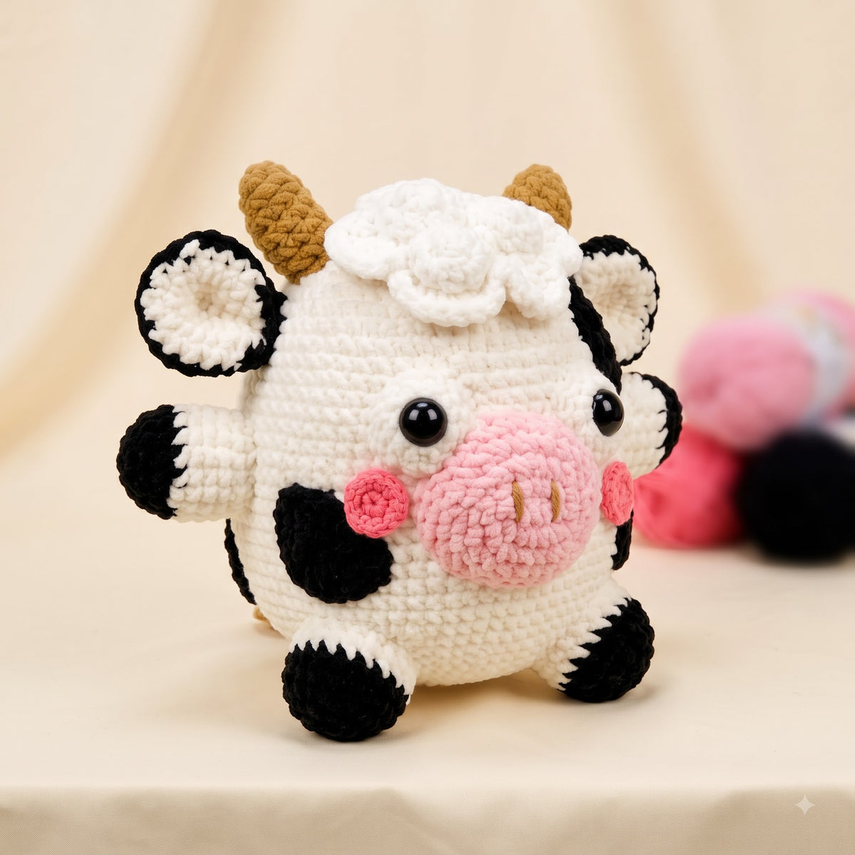 A crocheted plush cow toy with a white body, black spots, brown horns, and a pink snout, photographed on a soft beige fabric background with other plush toys blurred in the background.