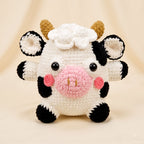 A crocheted plush cow toy with a white body, black spots, a pink snout and cheeks, small tan horns, and a white flower on its head, photographed against a soft cream background.
