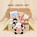 A crocheted cow plush toy sits in front of an open cardboard gift box filled with crochet yarn balls, crochet hooks, thread, and a printed instruction card. The text 'MAKE • CREATE • GIFT' appears above the box.