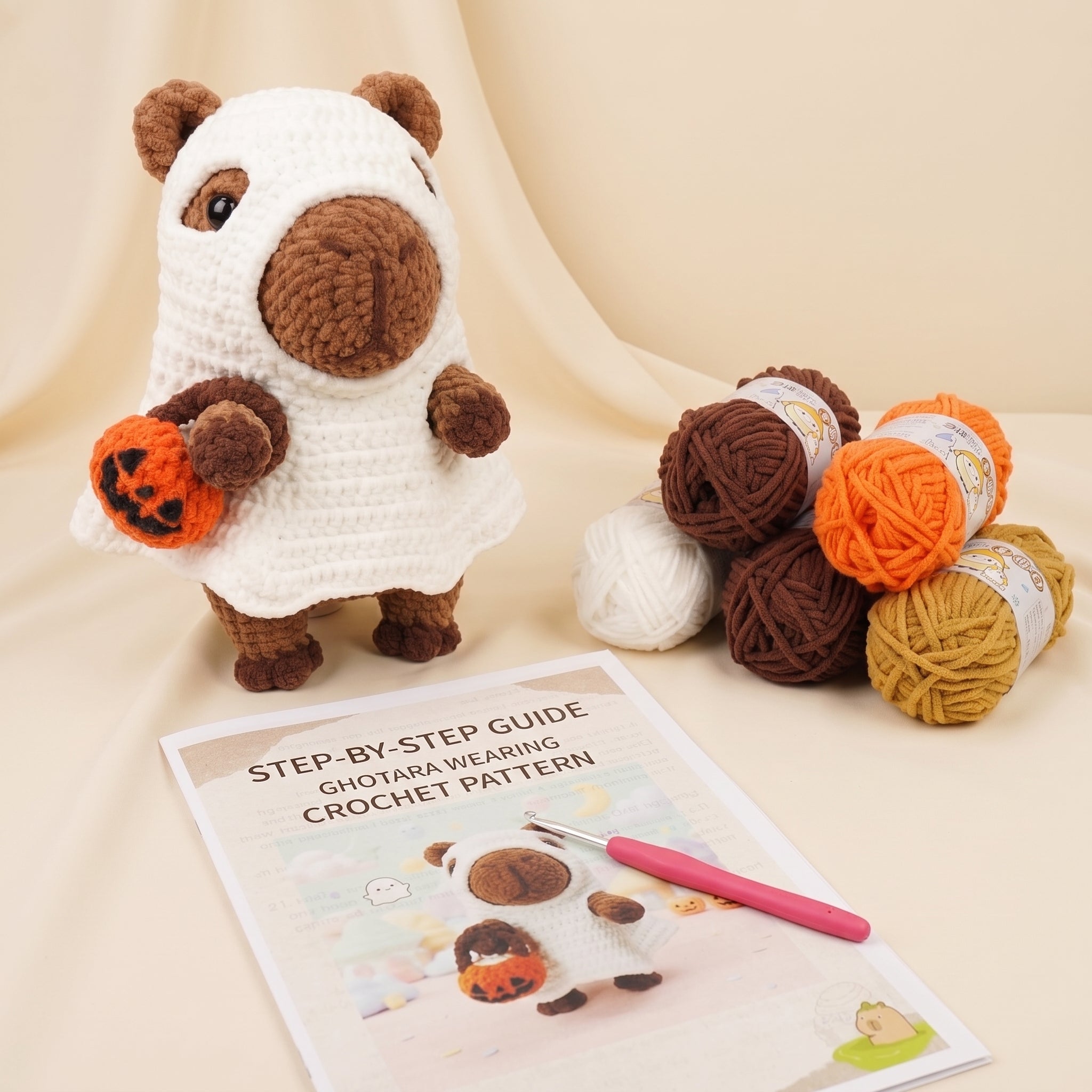 Crochet amigurumi ghost bear toy holding an orange pumpkin, displayed with a crochet pattern booklet, pink crochet hook, and multiple balls of yarn on a beige draped fabric background.