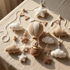 A crocheted baby mobile featuring a hot air balloon with a teddy bear, surrounded by clouds, stars, and wooden beads, displayed on a beige fabric surface.
