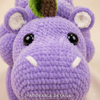 Close-up of a crocheted purple hippo plush toy with black bead eyes, white tusks, a green and brown horn, and white inner ears, on a light beige background with the text 'ADORABLE DETAILS' at the bottom.