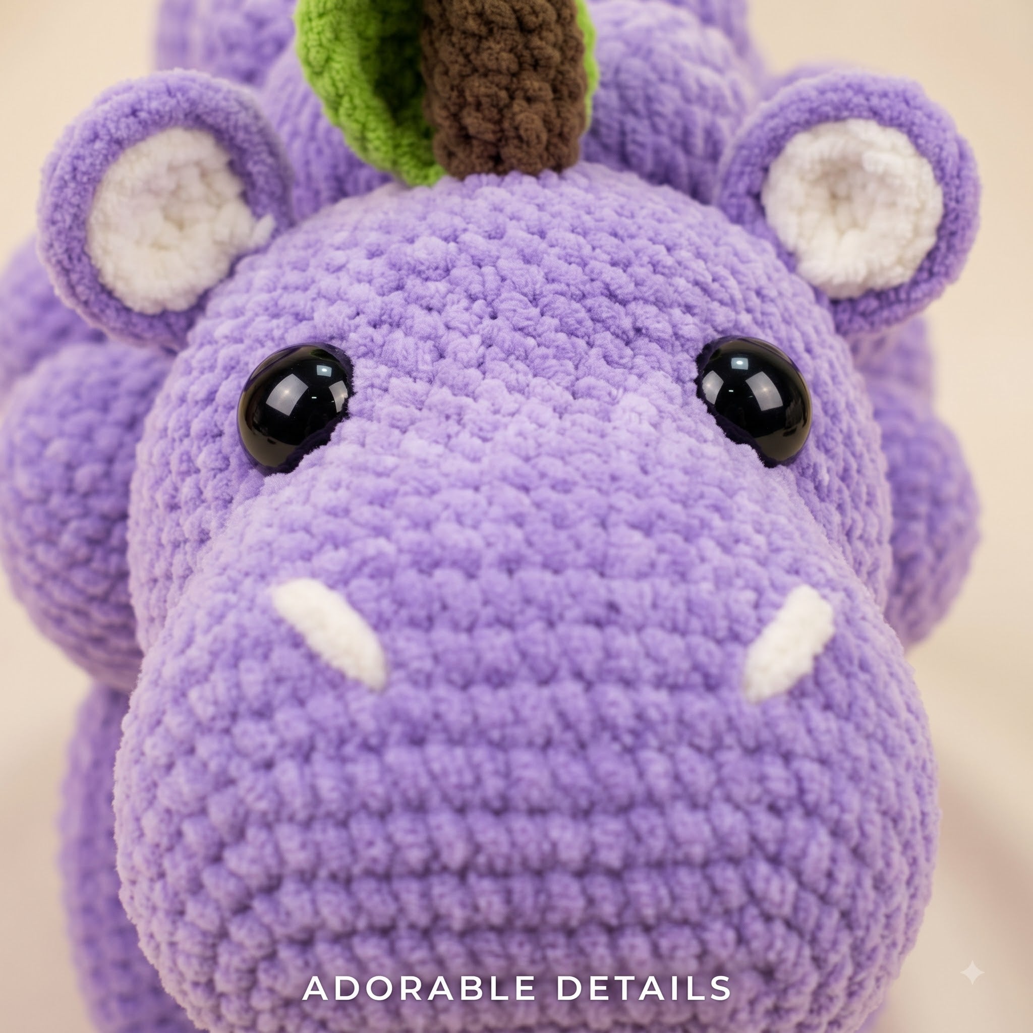 Close-up of a crocheted purple hippo plush toy with black bead eyes, white tusks, a green and brown horn, and white inner ears, on a light beige background with the text 'ADORABLE DETAILS' at the bottom.